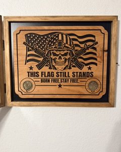Wooden shadow box plaque with an American flag, helmeted skull, crossed rifles, two coins and This Flag Still Stands text.